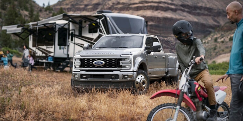 A silver 2024 Ford F-250 Super Duty parked near a trailer with a person riding a dirt bike to the right